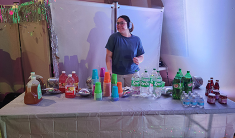 A young woman wears wireless headphones while standing behind a snack table.
