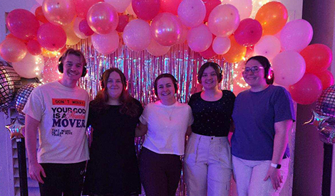 5 students stand in front of a balloon arch and smile while wearing headphones.