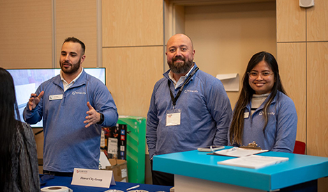 3 employees of Flower City Group stand behind a table. @ smile while 1 speaks with a student who stands off frame.
