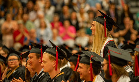 A graduate stands and smiles while other graduates sit and clap