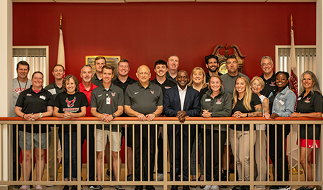 President Hayles and staff and coaches in the athletic department smile for a photo in the upper level of the Rinker Community Service Center.