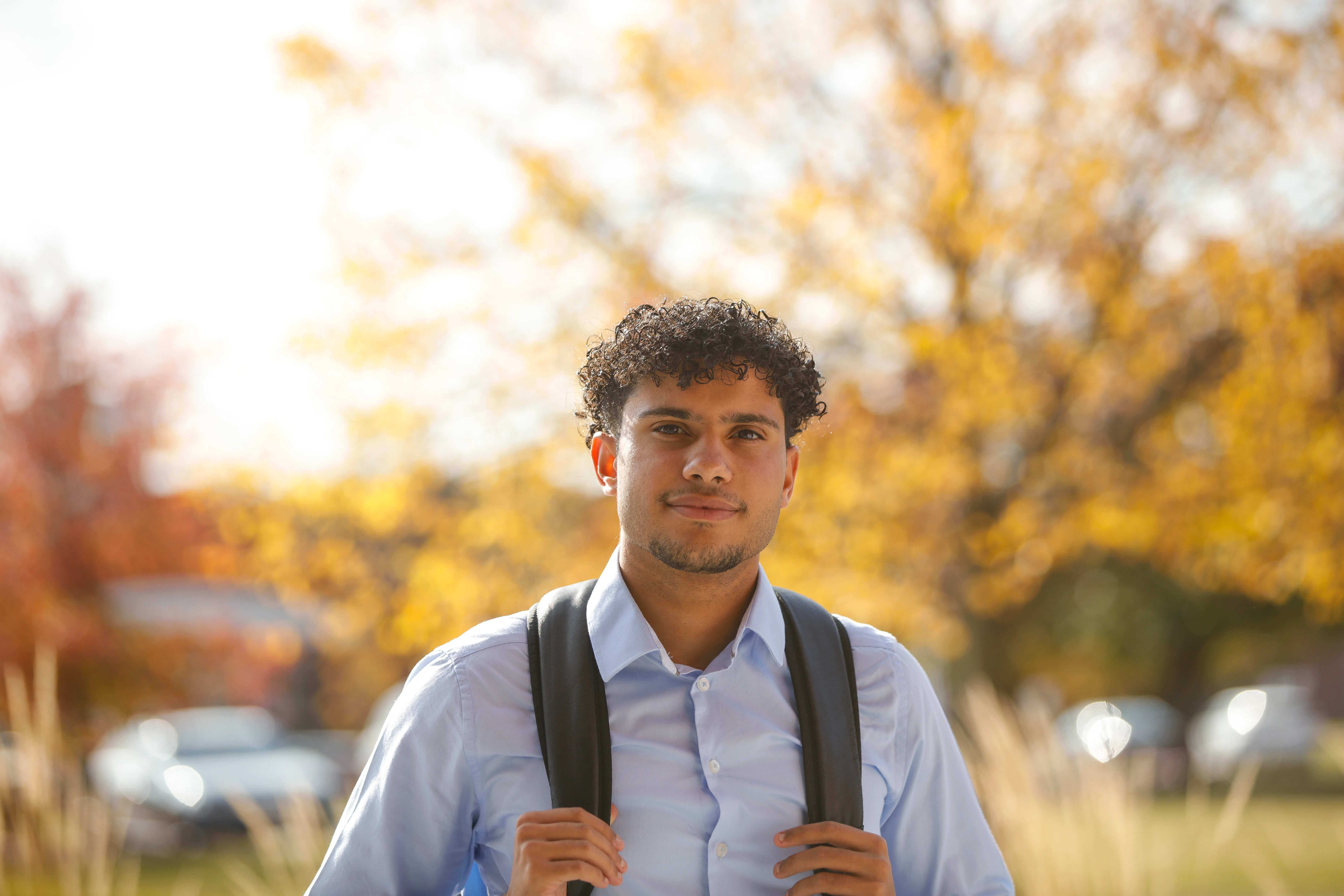 A young man smiles slightly in front of some trees with yellow leaves